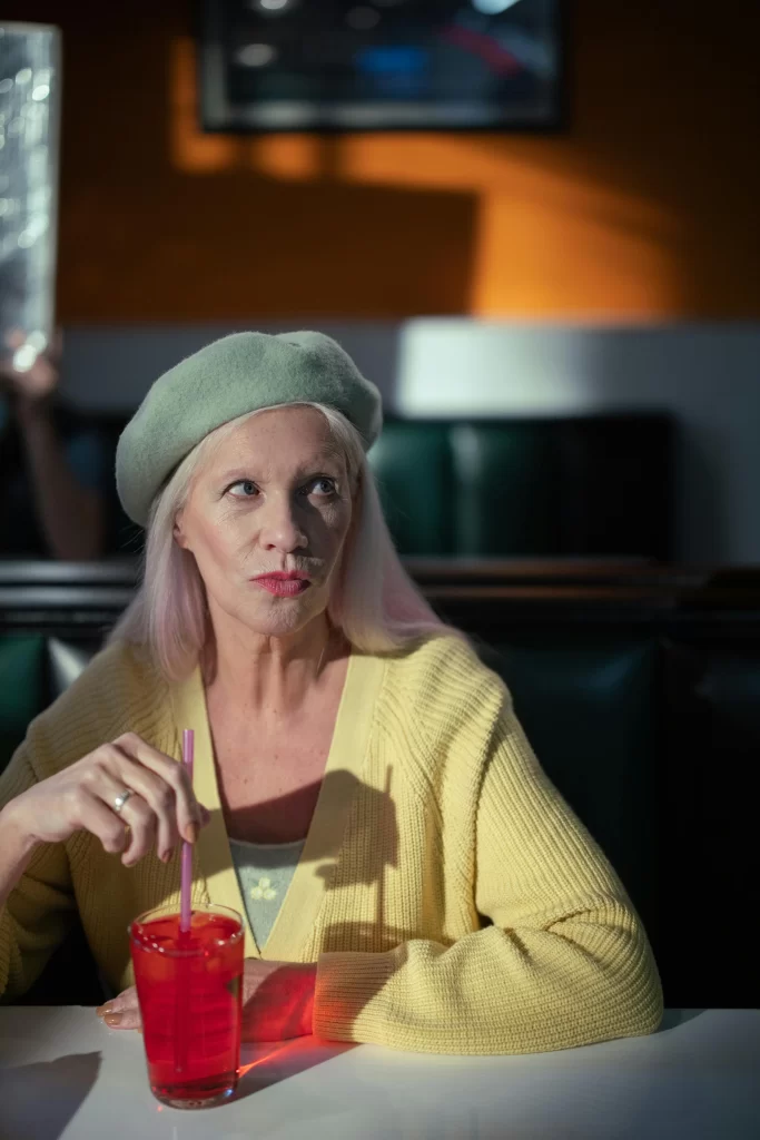 Older woman with long blonde hair wearing a yellow sweater and green beret, sitting in a diner while stirring a red drink with a straw.