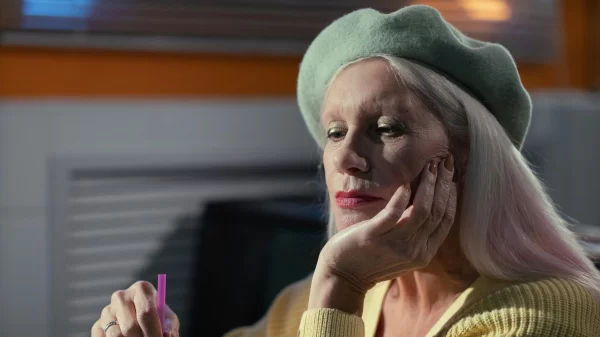 Older woman in a yellow sweater and green beret, resting her chin on her hand while stirring a red drink inside a retro-style diner.