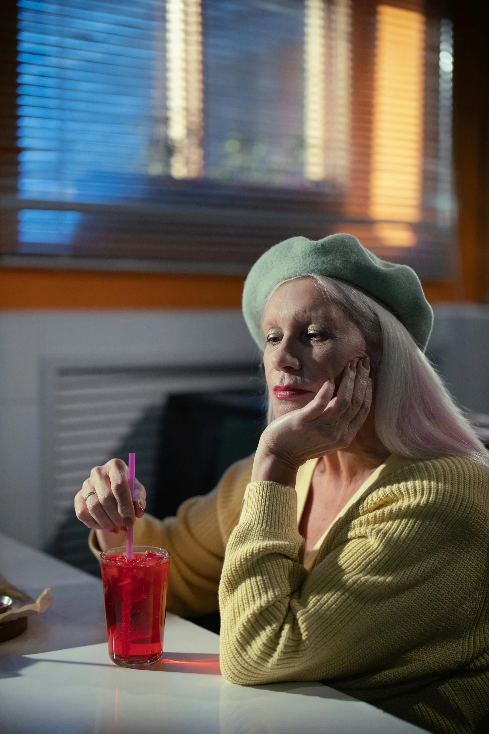 Older woman in a yellow sweater and green beret, resting her chin on her hand while stirring a red drink inside a retro-style diner.