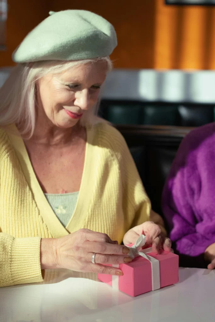 Smiling older woman wearing a yellow sweater and green beret, opening a small pink gift box at a diner table.