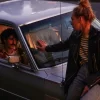 Woman sitting on a car while touching hands through the window with a man inside, dramatic evening lighting.