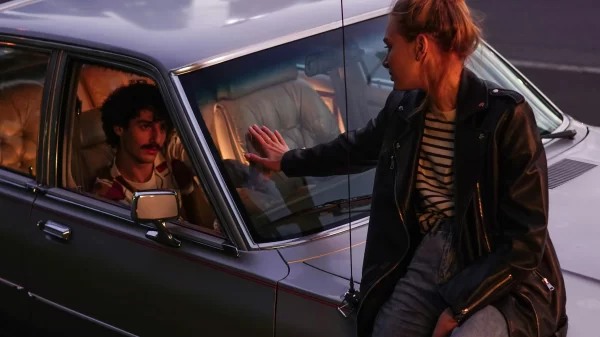 Woman sitting on a car while touching hands through the window with a man inside, dramatic evening lighting.