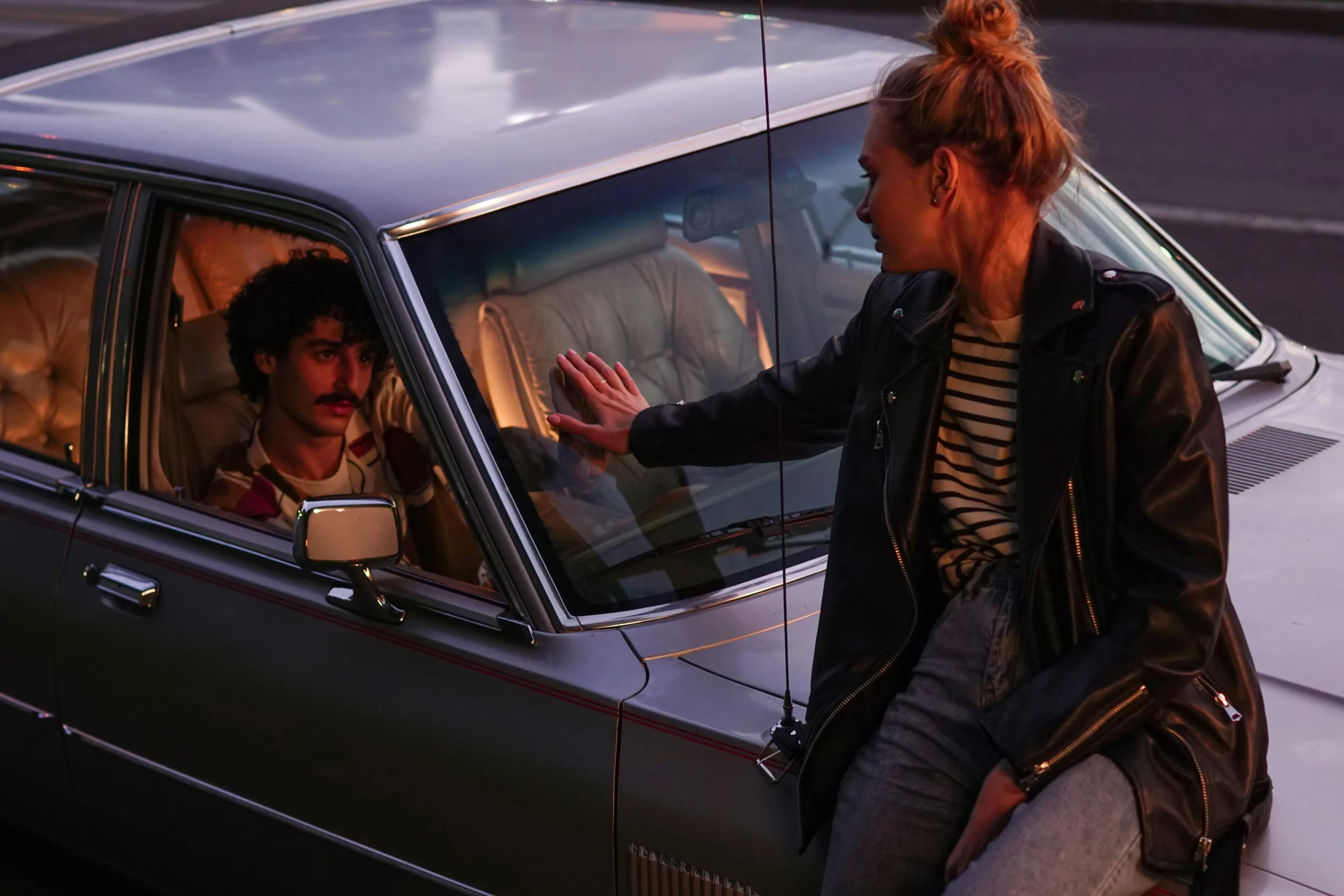 Woman sitting on a car while touching hands through the window with a man inside, dramatic evening lighting.