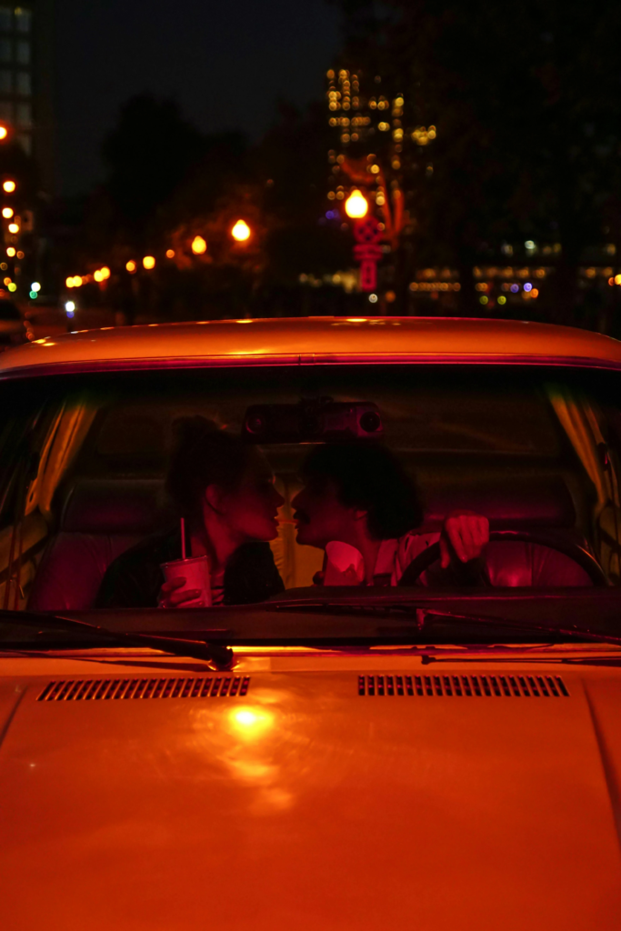 Couple sitting in a car at night, leaning in for a kiss under warm streetlight glow.