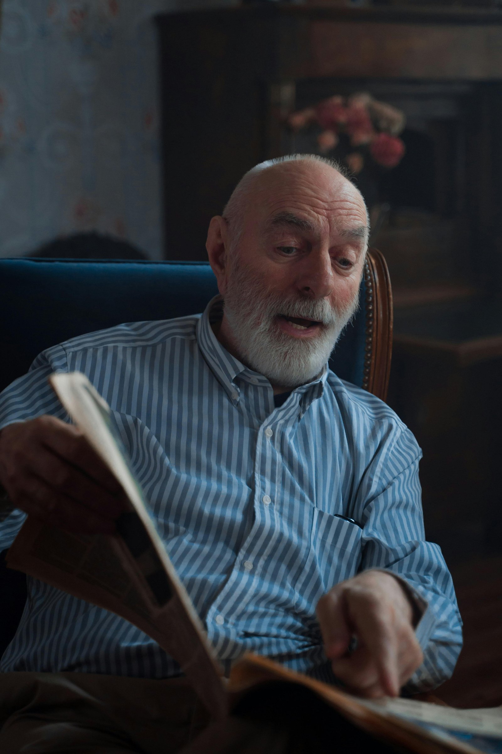Older man with a gray beard reading a newspaper while sitting in a cozy living room chair.
