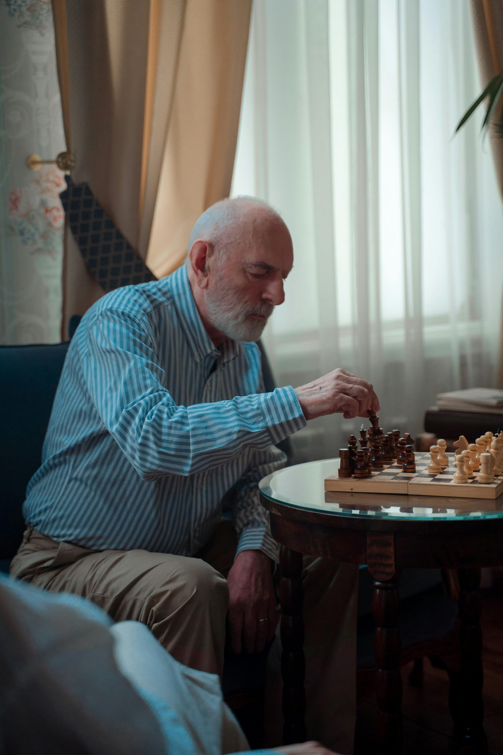 Older man with a white beard playing chess alone at a small round table in a softly lit living room.