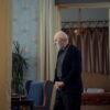 Elderly man with a white beard standing thoughtfully in a vintage-style living room with patterned wallpaper and curtains.