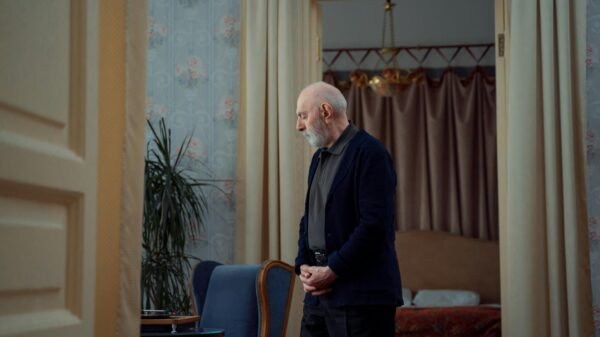 Elderly man with a white beard standing thoughtfully in a vintage-style living room with patterned wallpaper and curtains.
