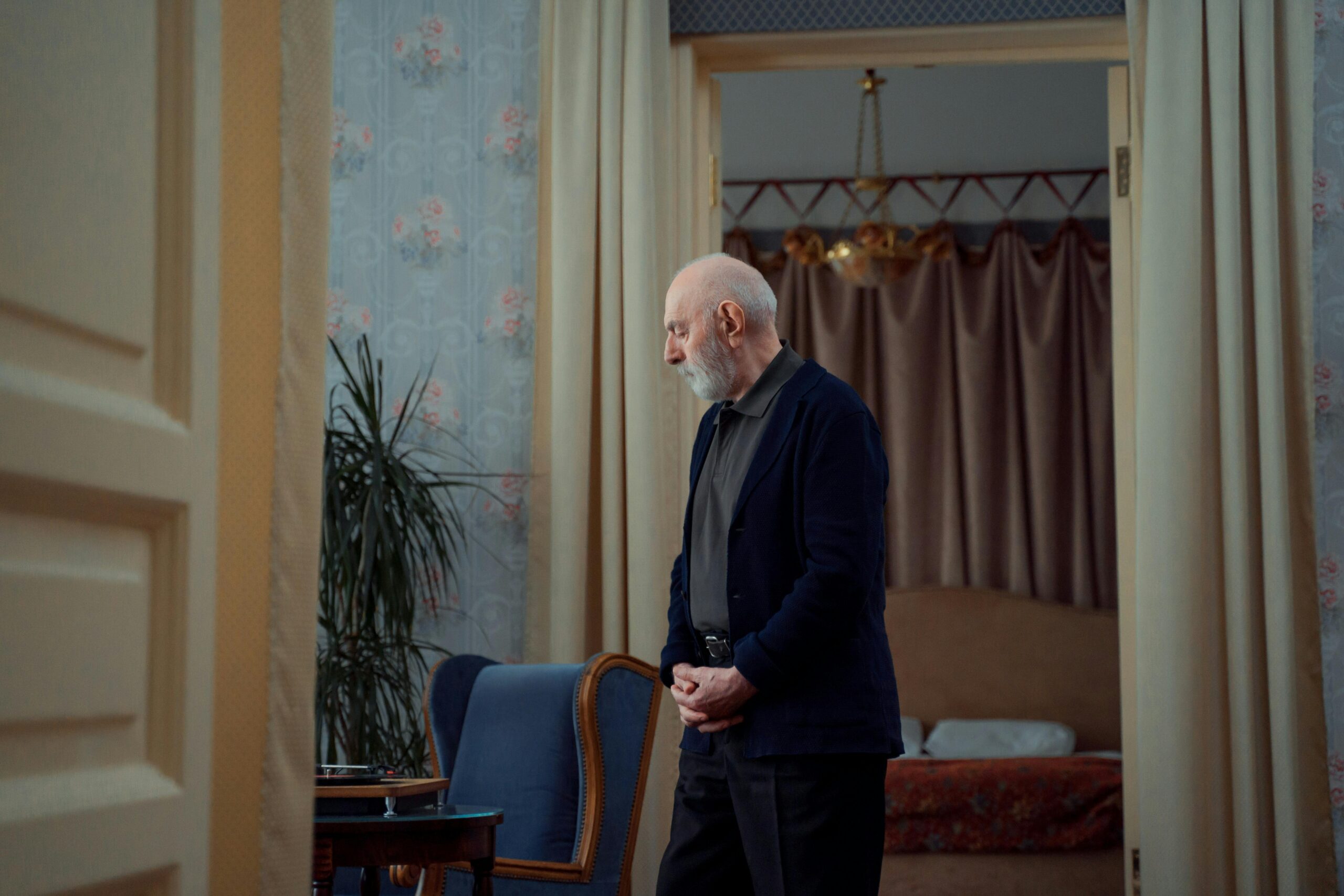 Elderly man with a white beard standing thoughtfully in a vintage-style living room with patterned wallpaper and curtains.
