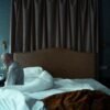 Older man sitting on the edge of his bed in a quiet bedroom, looking down in thought.