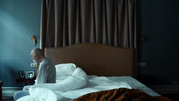 Older man sitting on the edge of his bed in a quiet bedroom, looking down in thought.