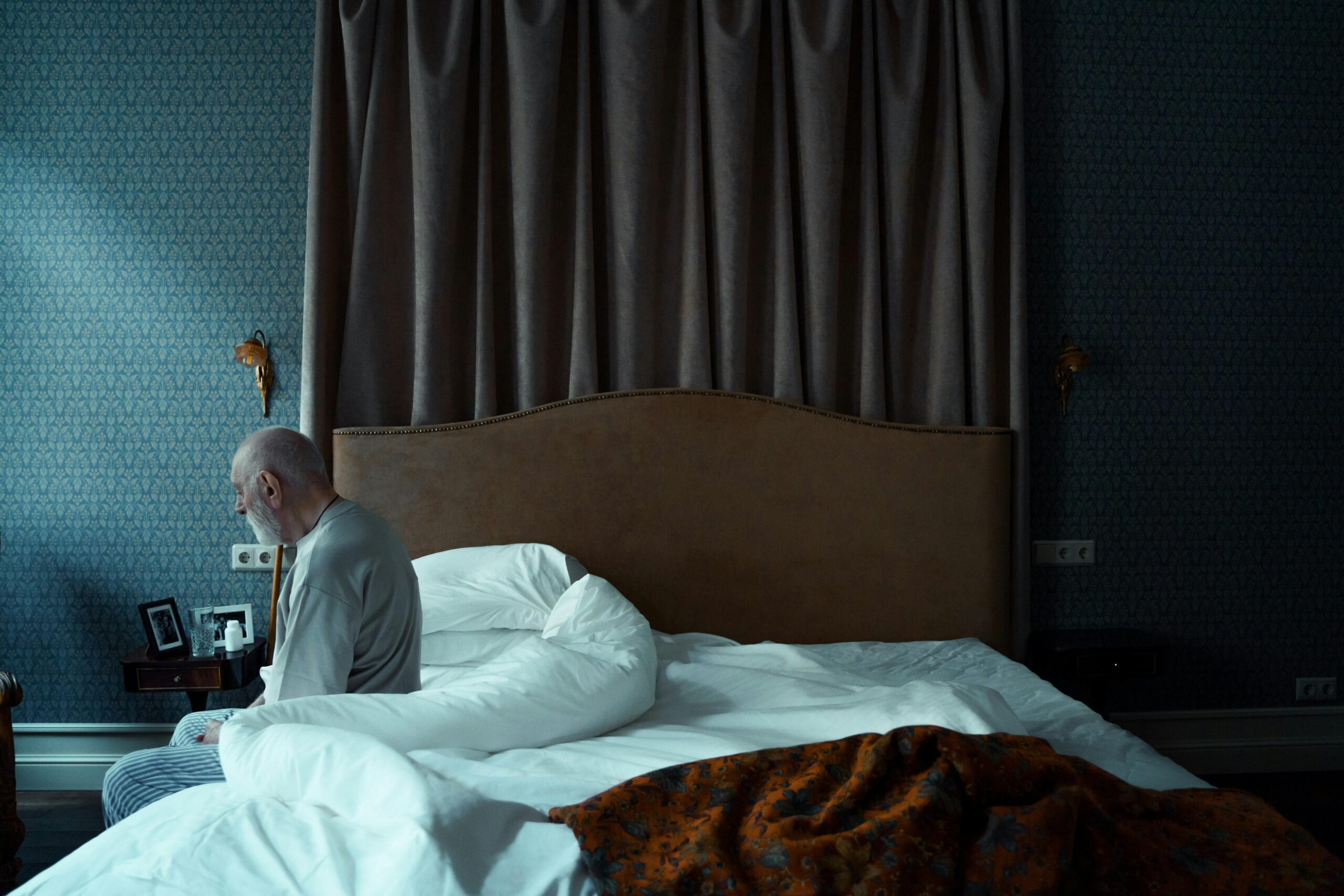Older man sitting on the edge of his bed in a quiet bedroom, looking down in thought.
