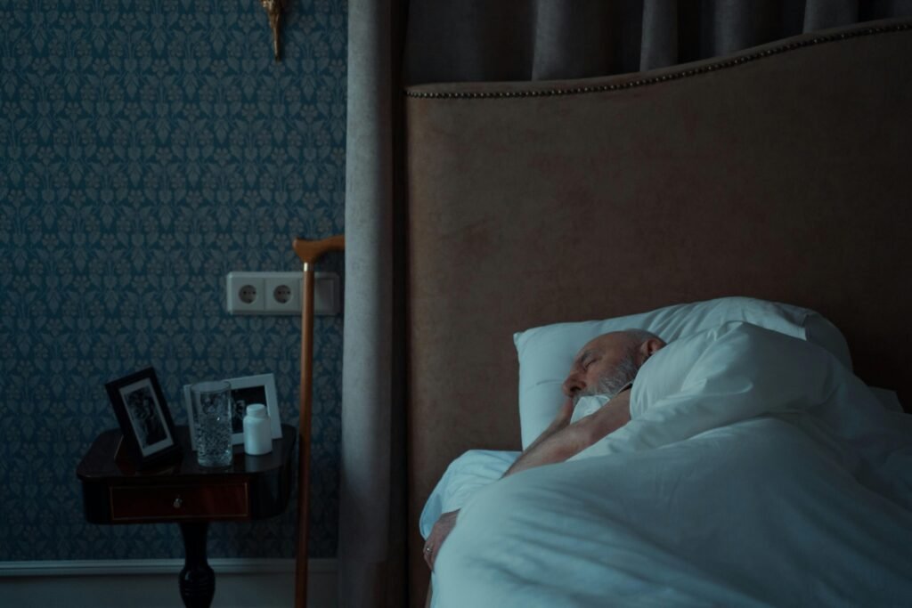 Older man sleeping in bed in a dim, peaceful bedroom with framed photos on the nightstand.