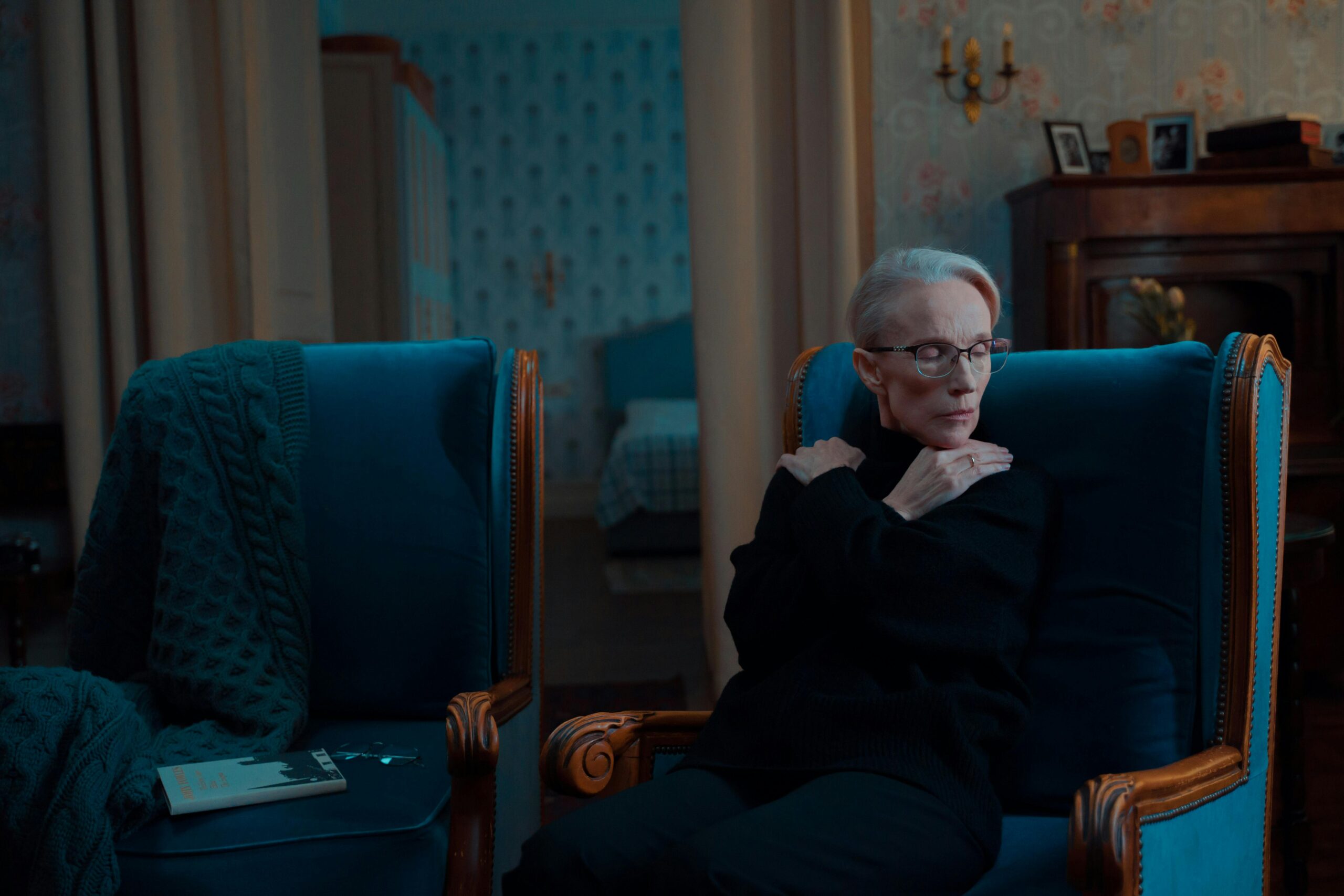 Older woman sitting alone in an armchair, reflecting on future plans in a quiet, dimly lit room