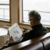 Elderly man reading a newspaper while sitting on a ferry, looking out toward the water.