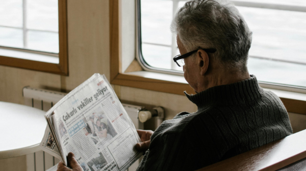 Elderly man reading a newspaper while sitting on a ferry, looking out toward the water.