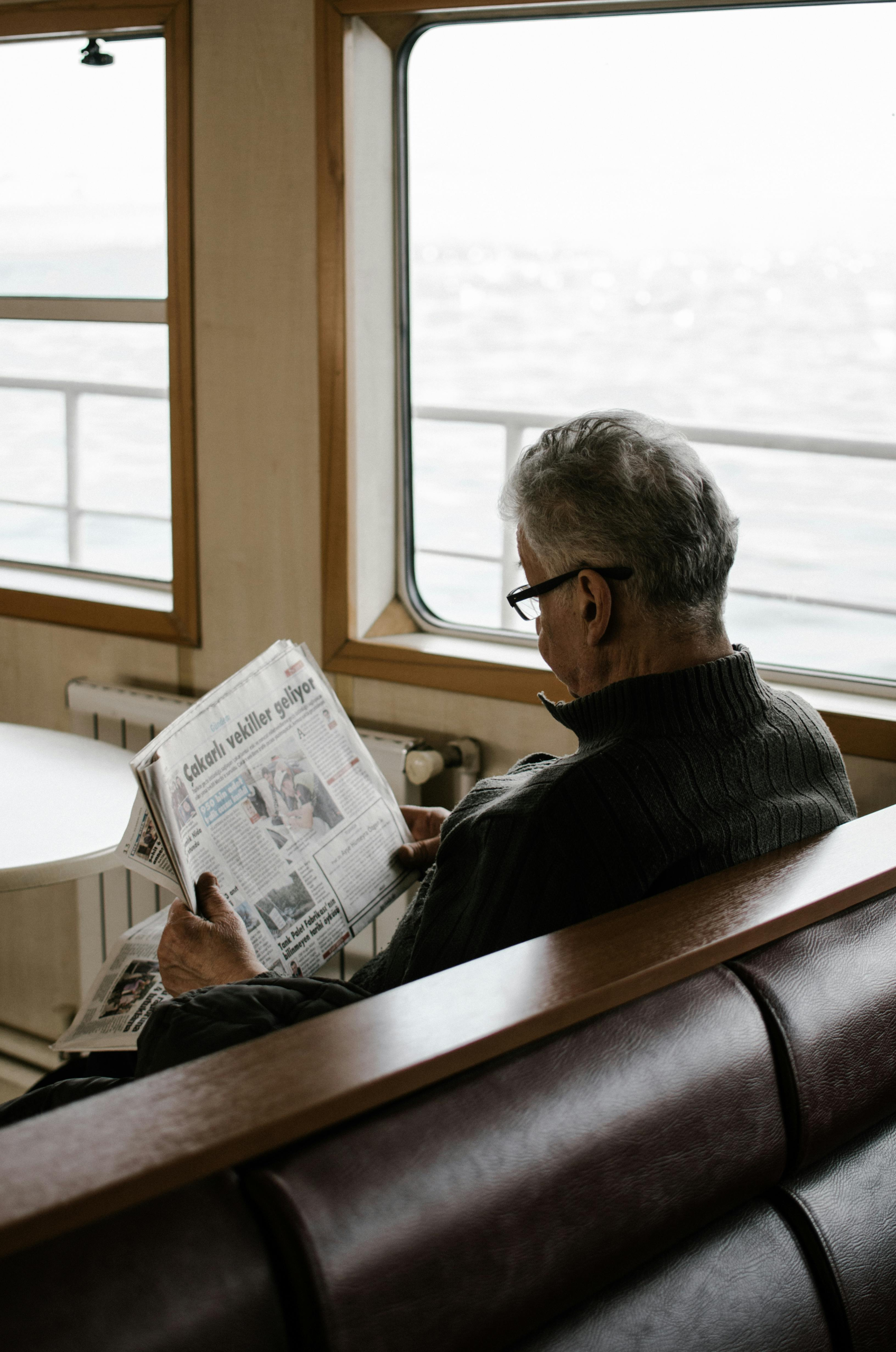 Elderly man reading a newspaper while sitting on a ferry, looking out toward the water.