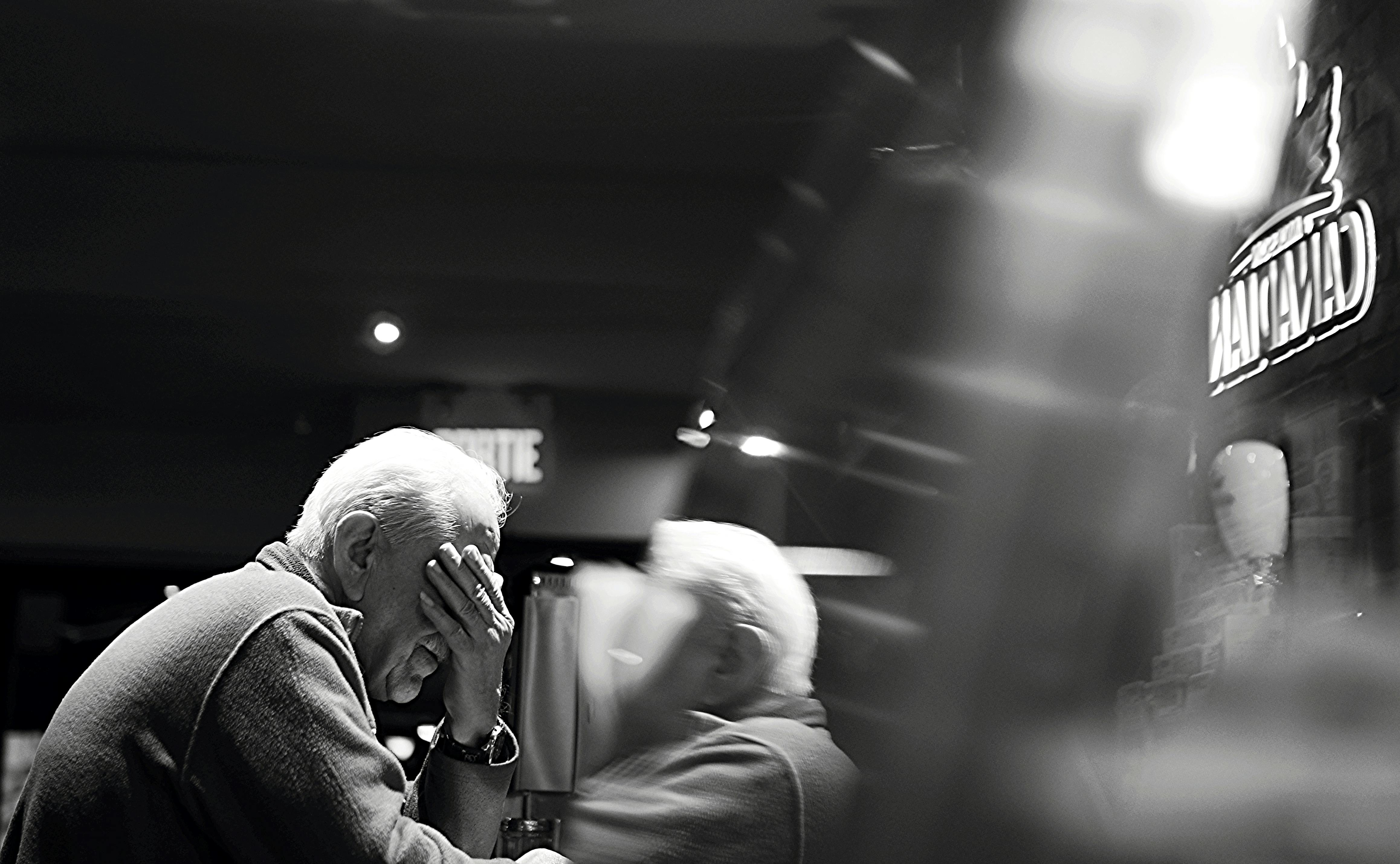Older man sitting alone in a bar with his hand on his face, looking stressed or overwhelmed.