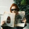 Person with red hair wearing sunglasses, reading a vintage LIFE magazine featuring Picasso while seated indoors near greenery