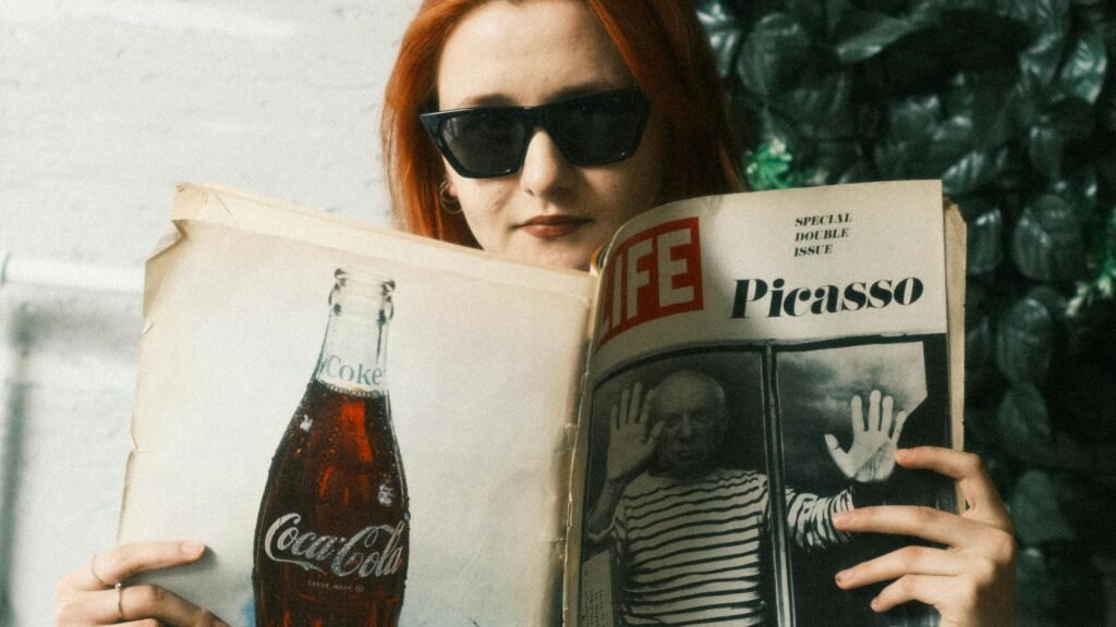 Person with red hair wearing sunglasses, reading a vintage LIFE magazine featuring Picasso while seated indoors near greenery