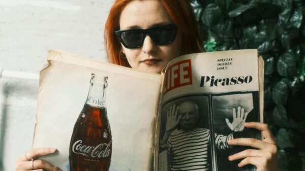 Person with red hair wearing sunglasses, reading a vintage LIFE magazine featuring Picasso while seated indoors near greenery
