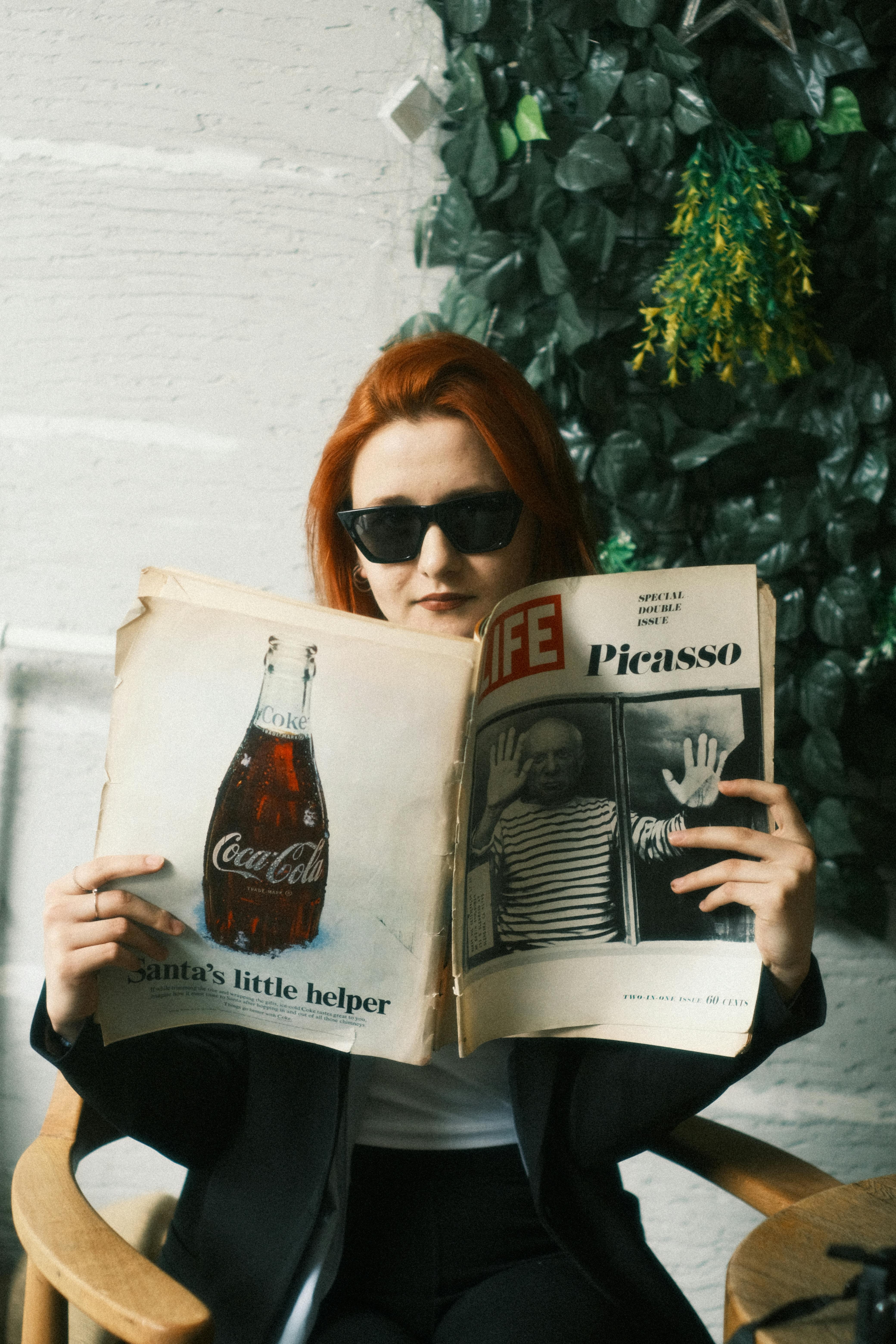 Person with red hair wearing sunglasses, reading a vintage LIFE magazine featuring Picasso while seated indoors near greenery