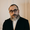 Middle-aged man with beard and glasses standing calmly in a dark suit, expressing quiet reflection