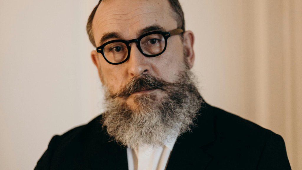 Middle-aged man with beard and glasses standing calmly in a dark suit, expressing quiet reflection