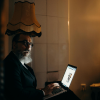Older man with a beard working on a laptop in a softly lit room at night