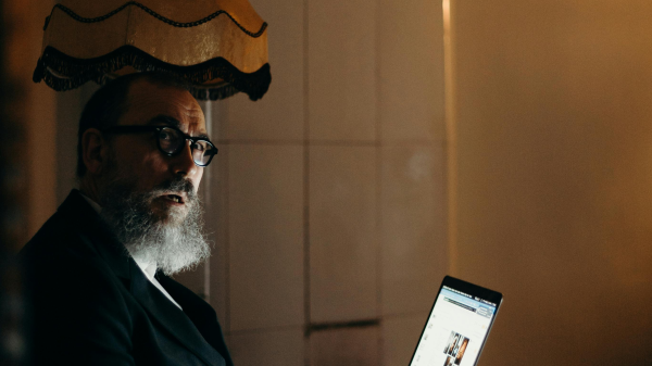 Older man with a beard working on a laptop in a softly lit room at night