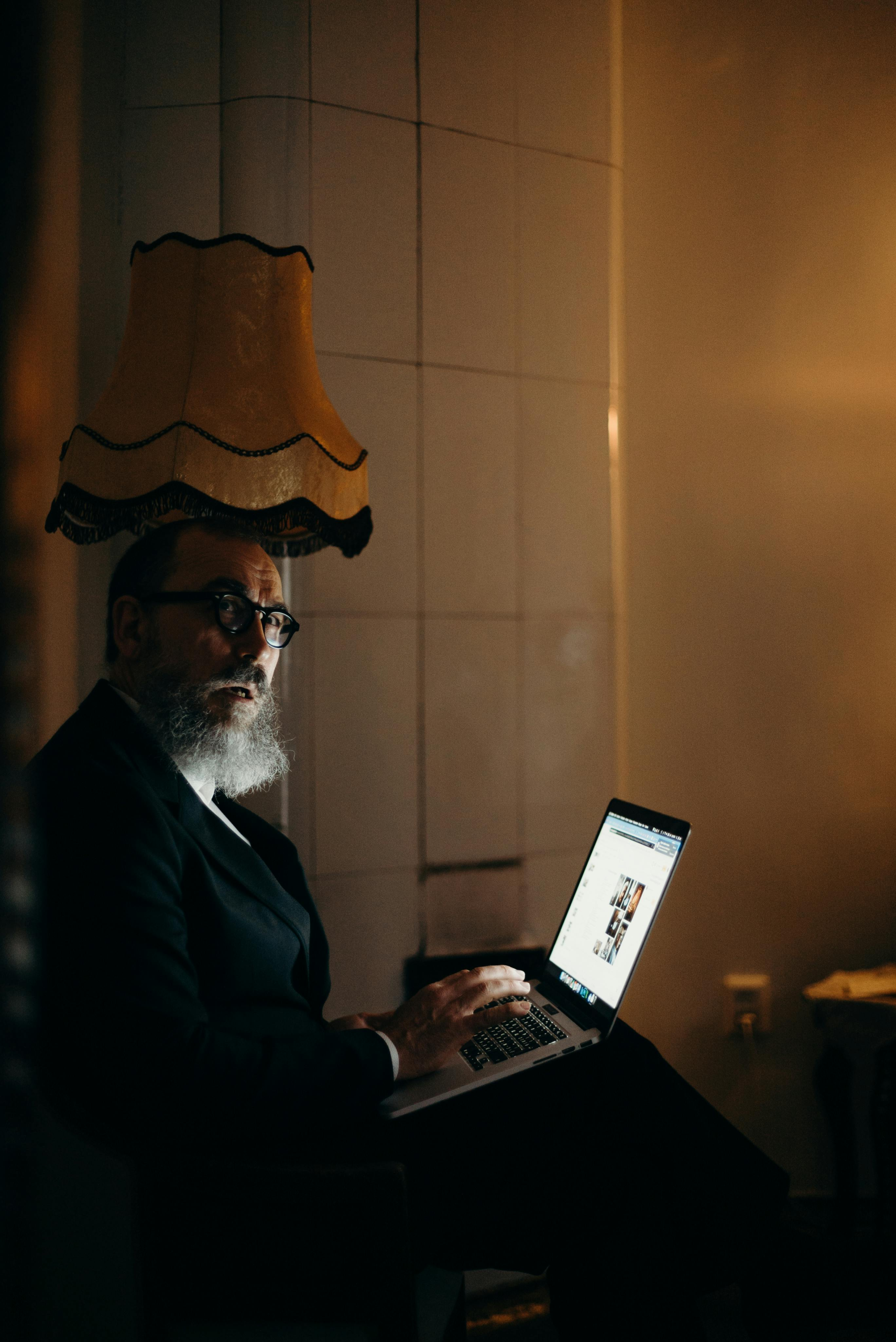 Older man with a beard working on a laptop in a softly lit room at night