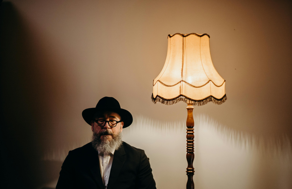 Bearded man in a hat sitting beside a warm lamp, symbolizing introspection and personal transformation