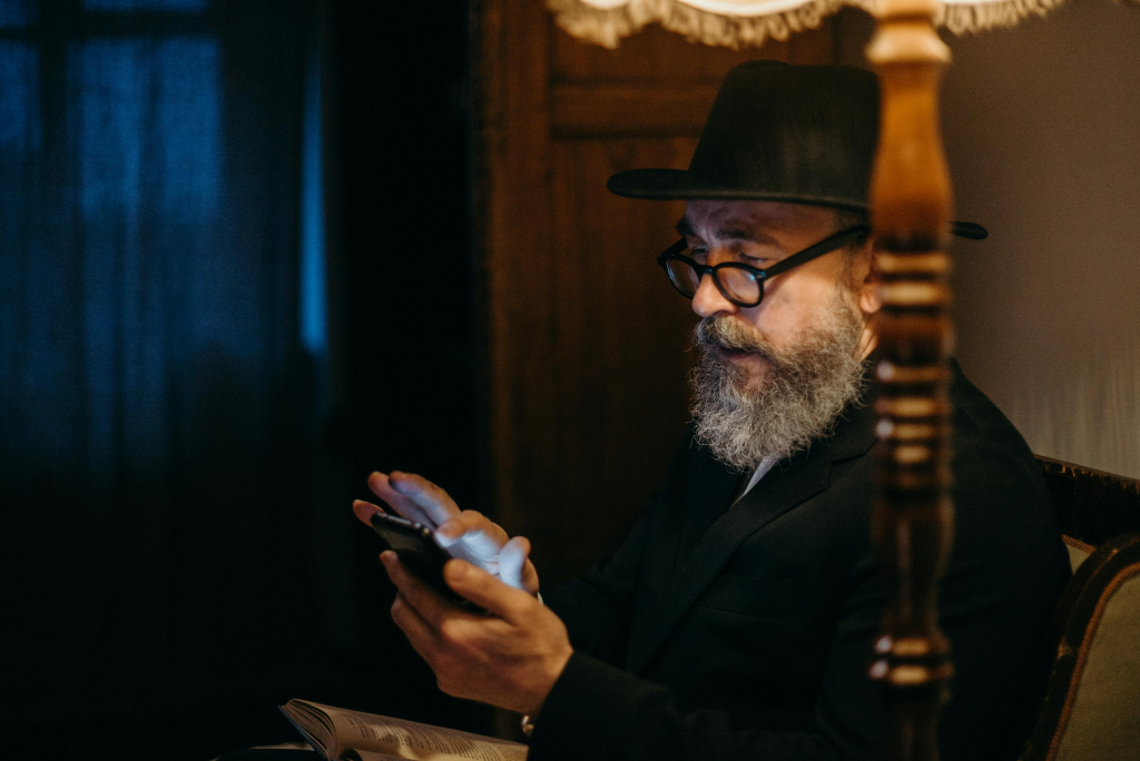 Older man with a gray beard using a smartphone while sitting indoors under warm lighting