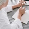 Older woman sitting at a table writing in a notebook beside a laptop, reflecting or planning in a quiet home setting
