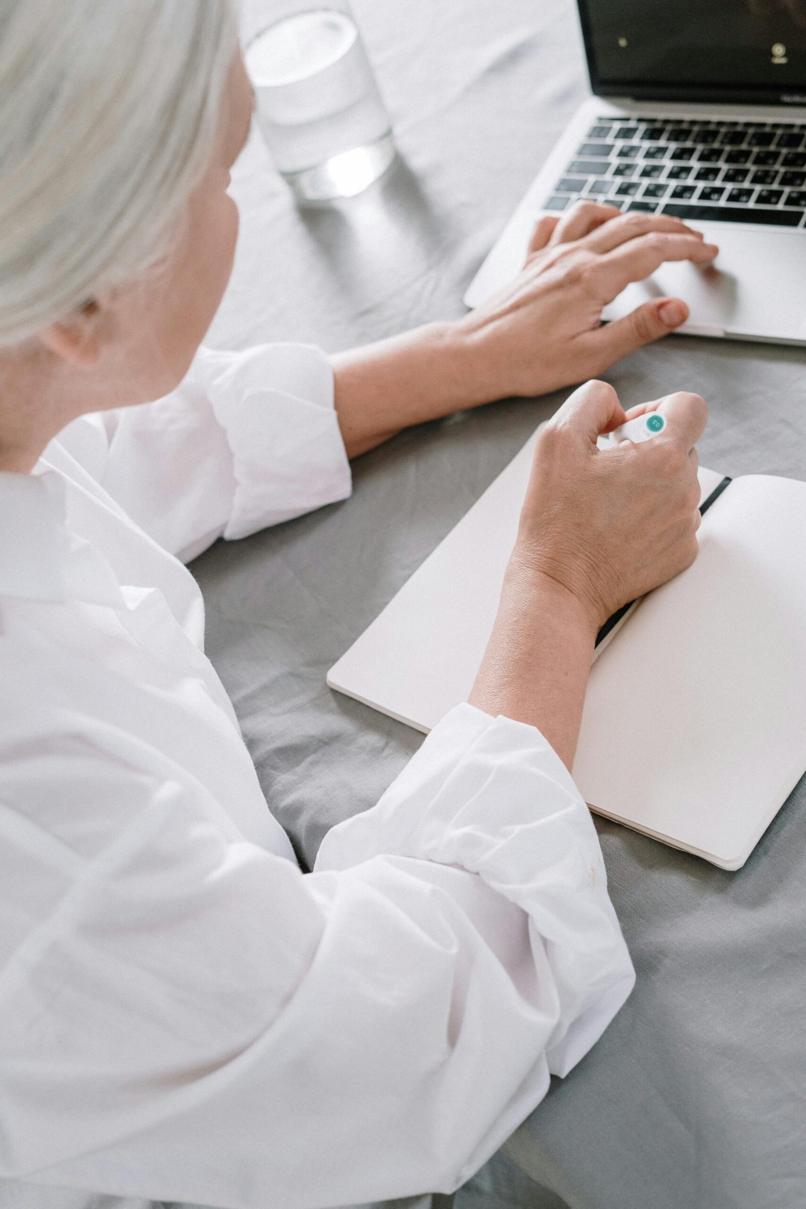 Older woman sitting at a table writing in a notebook beside a laptop, reflecting or planning in a quiet home setting