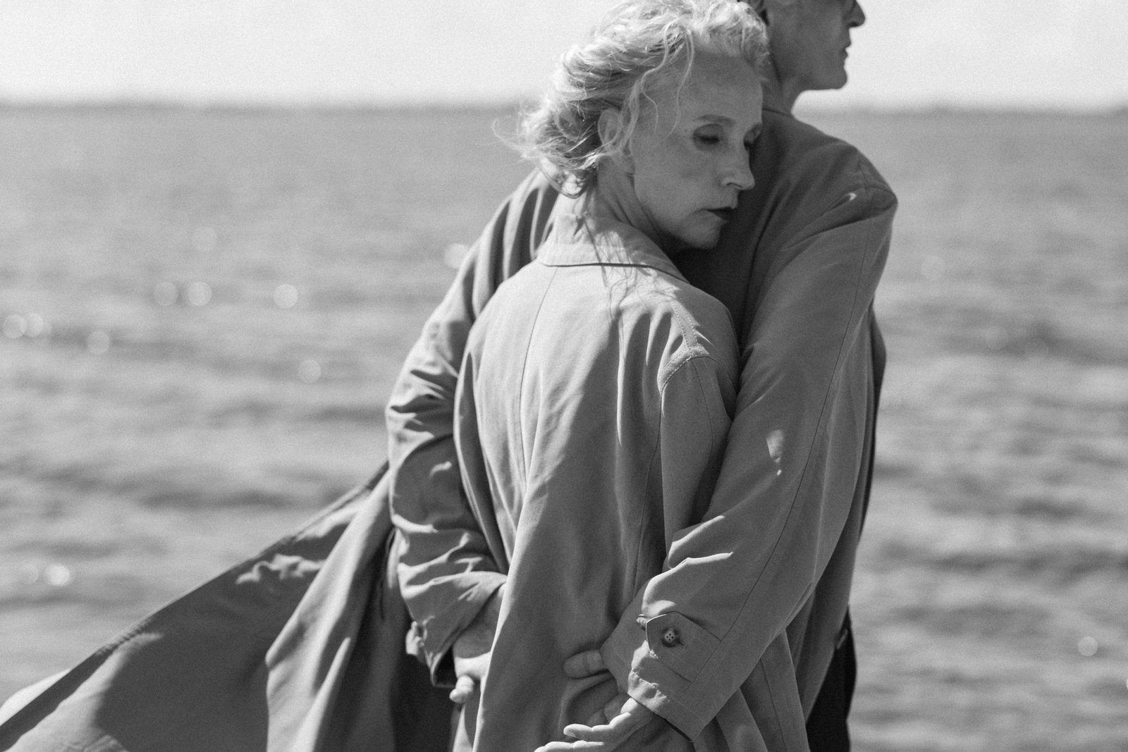 Two older adults standing by the ocean, holding each other closely in a quiet, intimate moment