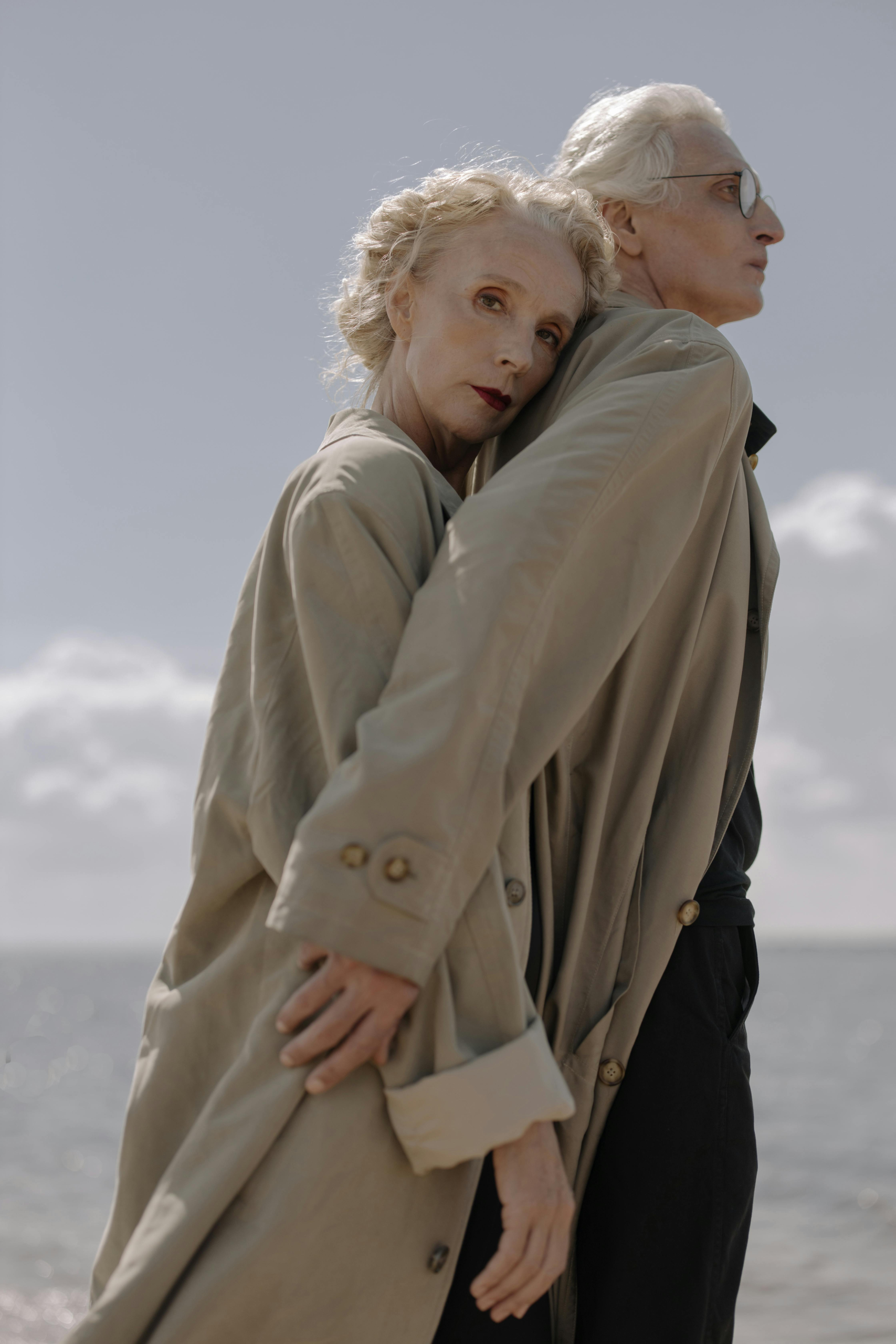 Older couple embracing near the ocean, wearing classic coats and looking thoughtful