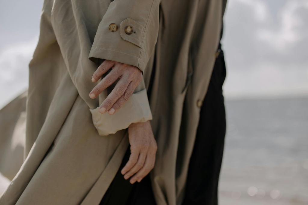 Close-up of two middle-aged adults wearing neutral trench coats, hands relaxed by the seaside