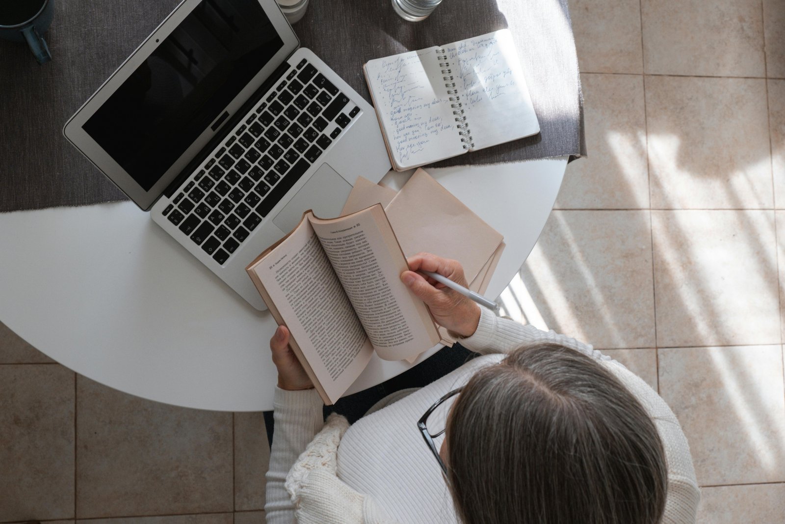 Middle-aged woman reading a book at a desk with a laptop and notebook, reflecting on learning and simplicity in midlife