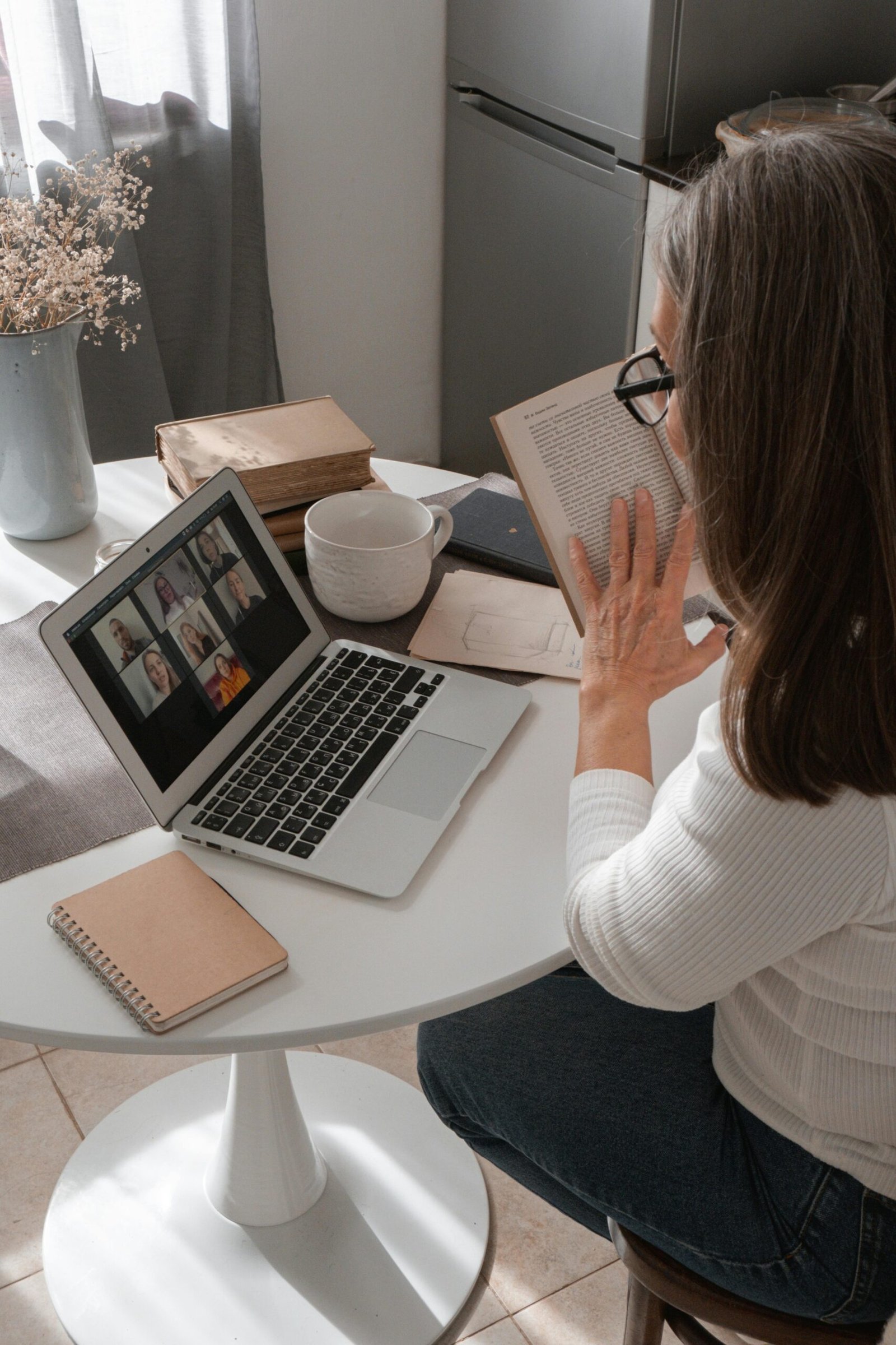 Woman in her 50s reading a book while attending a video call at home, balancing fewer tools and intentional work
