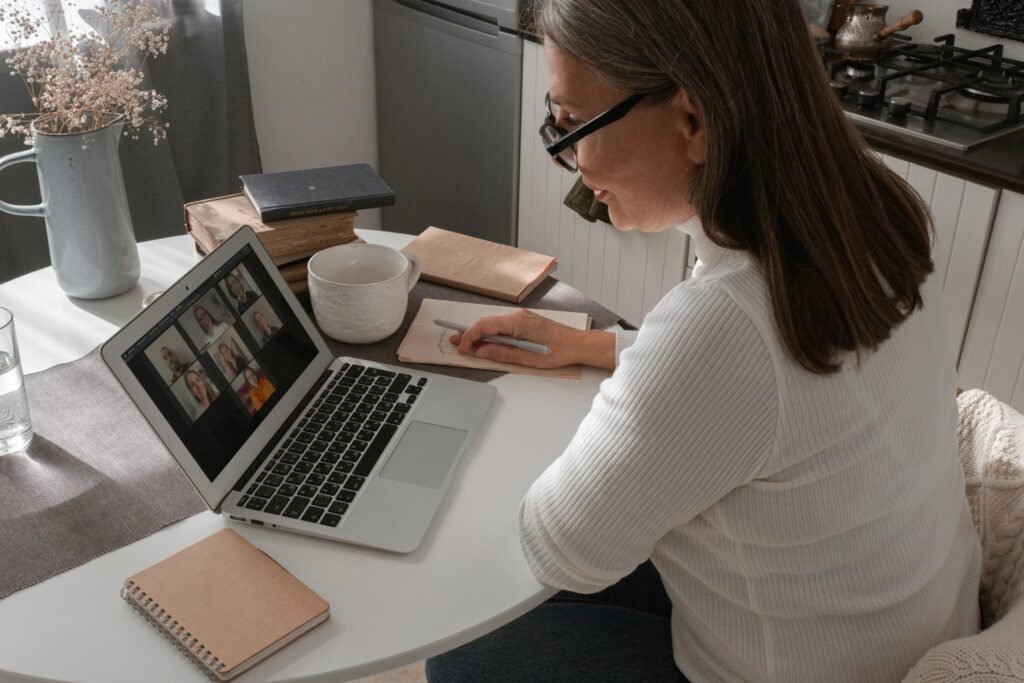 Woman in her 60s attending an online video meeting from home, taking notes during a virtual discussion