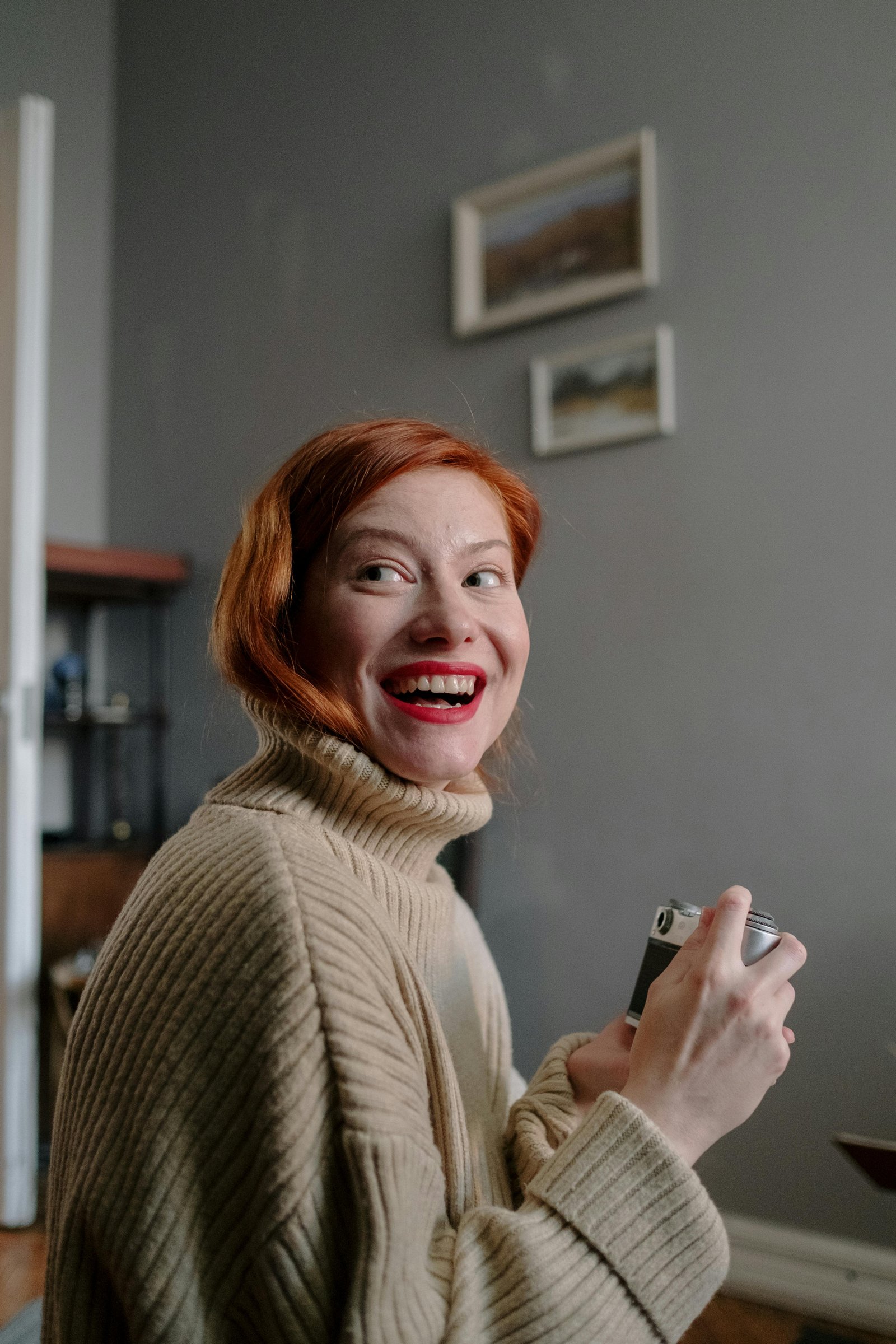 Smiling red-haired woman in a cozy sweater holding a camera indoors, expressing confidence and warmth