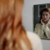 Red-haired woman looking at her reflection in a mirror, hands on face, thoughtful and self-aware