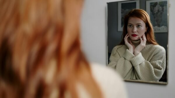 Red-haired woman looking at her reflection in a mirror, hands on face, thoughtful and self-aware