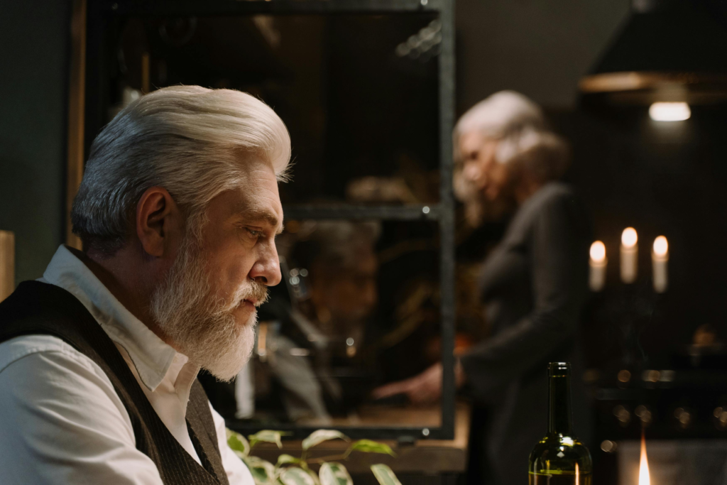 Older man with gray hair sitting thoughtfully at a candlelit table, reflecting in a mirror during an intimate evening at home