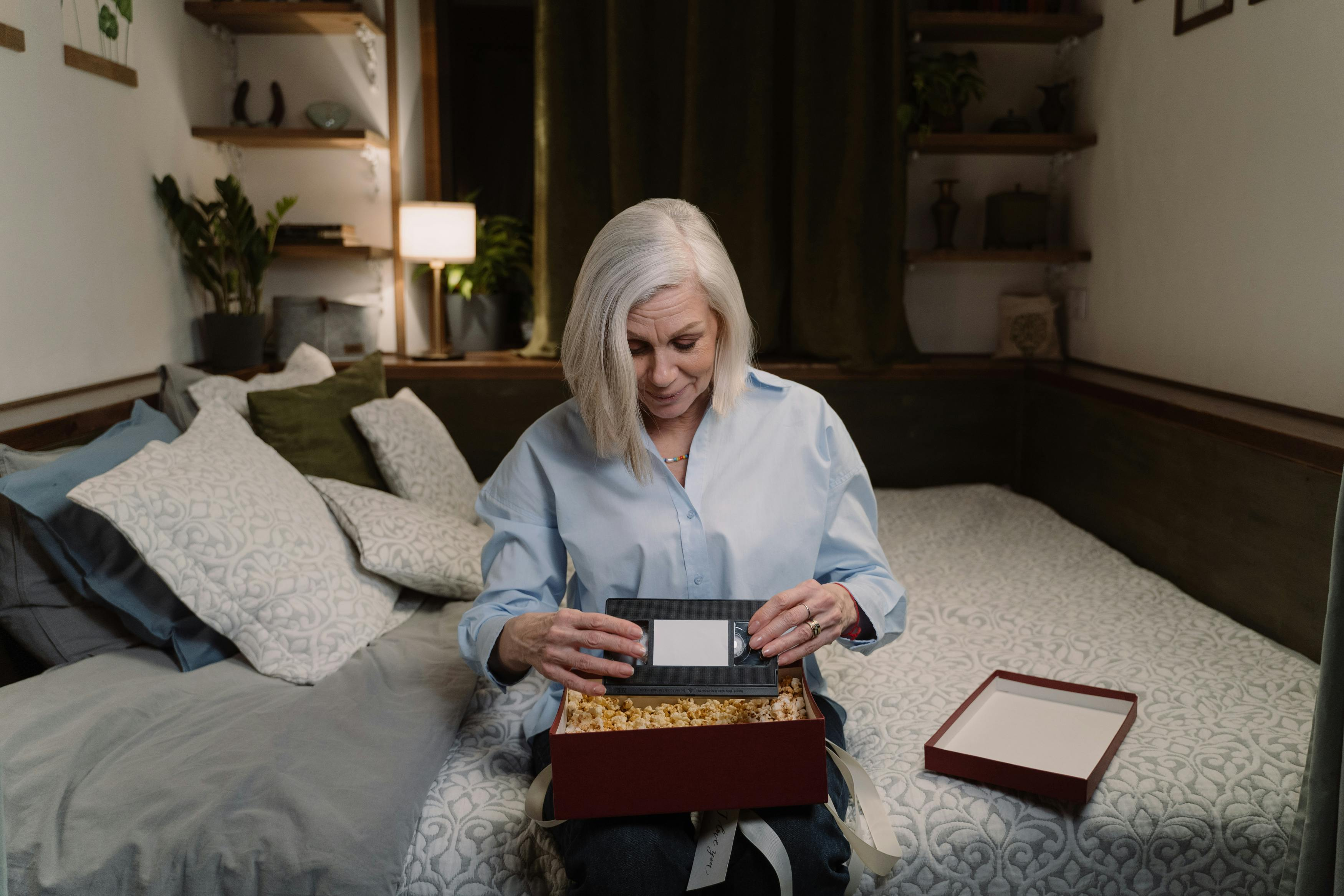 Older woman sitting on her bed opening a memory box filled with popcorn and a VHS tape, reflecting quietly at home
