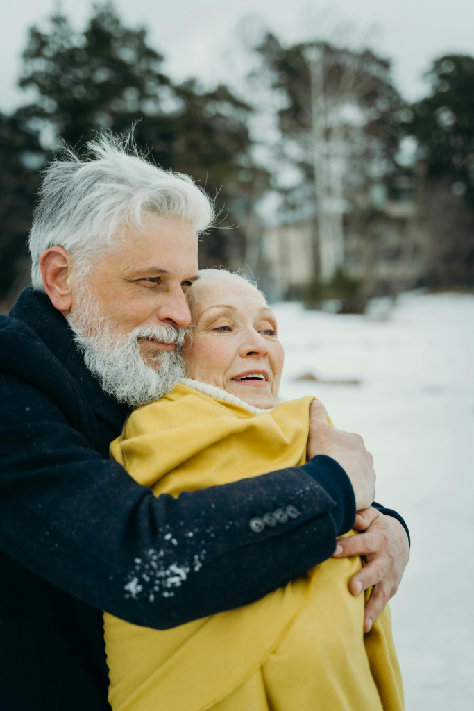Older couple standing together under bare winter trees, sharing a quiet, intimate moment outdoors