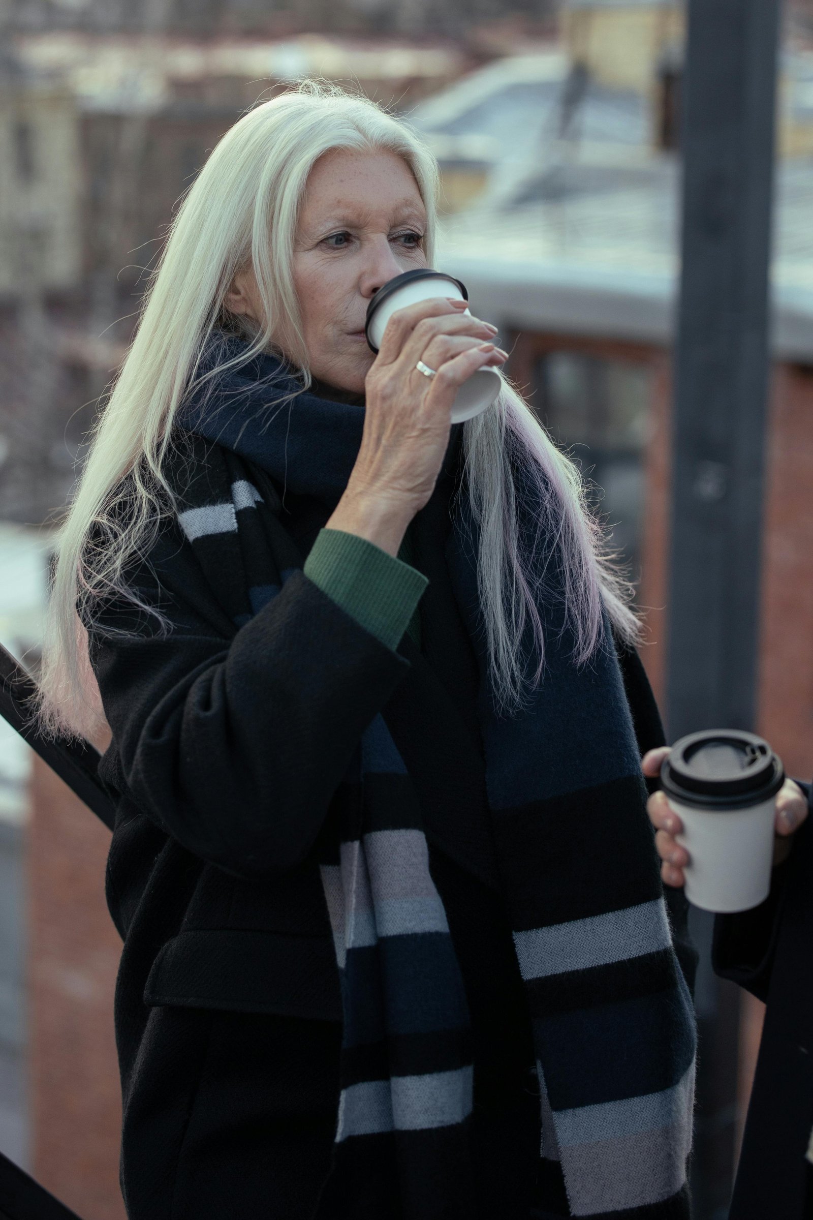 Older woman standing outdoors holding a takeaway coffee, pausing quietly and reflecting during a calm moment in her day