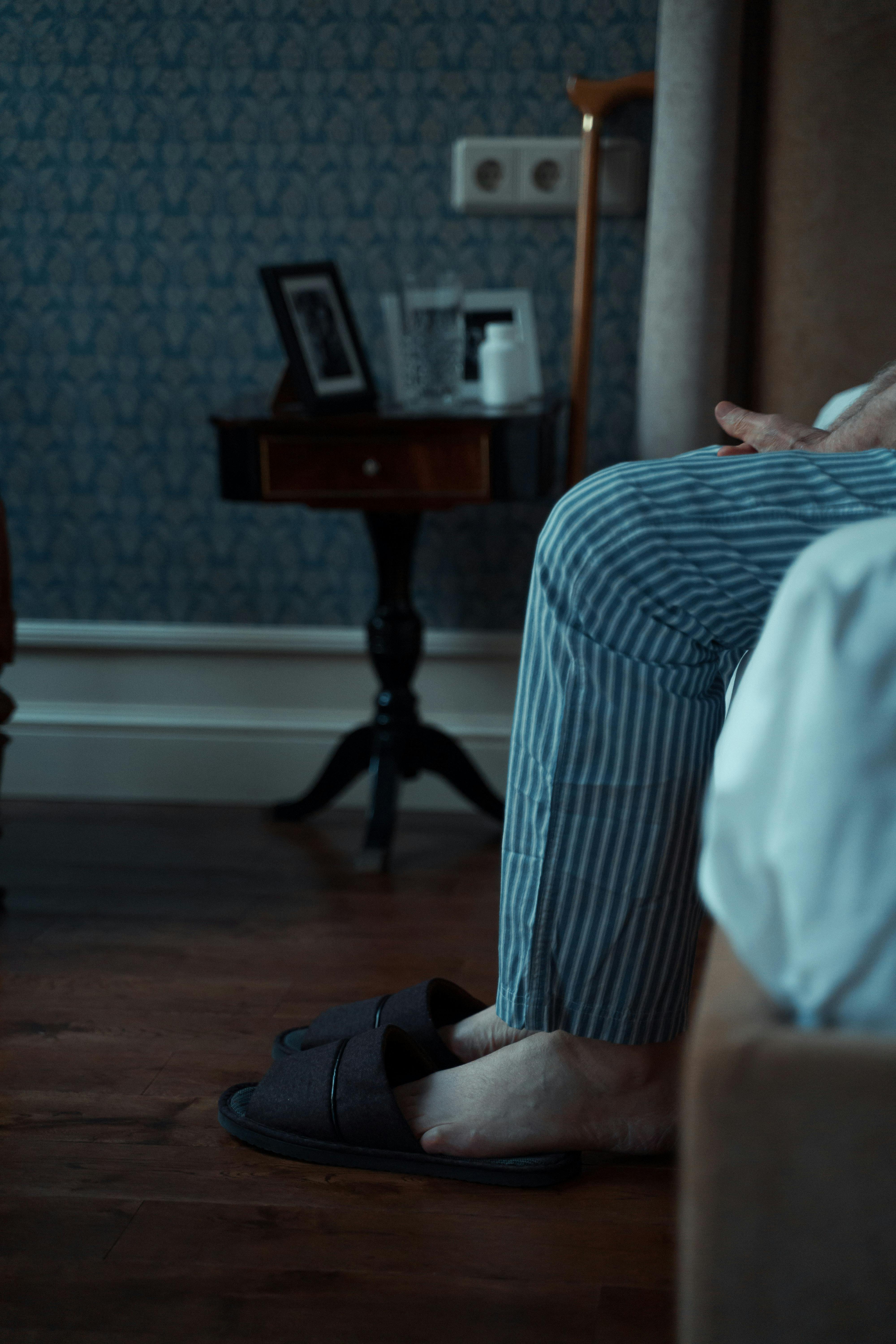 Older man sitting quietly at home, feet in slippers, reflecting in a calm, dimly lit room