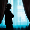 Silhouette of an older woman standing by a window, holding her arms close while thinking about a midlife shift